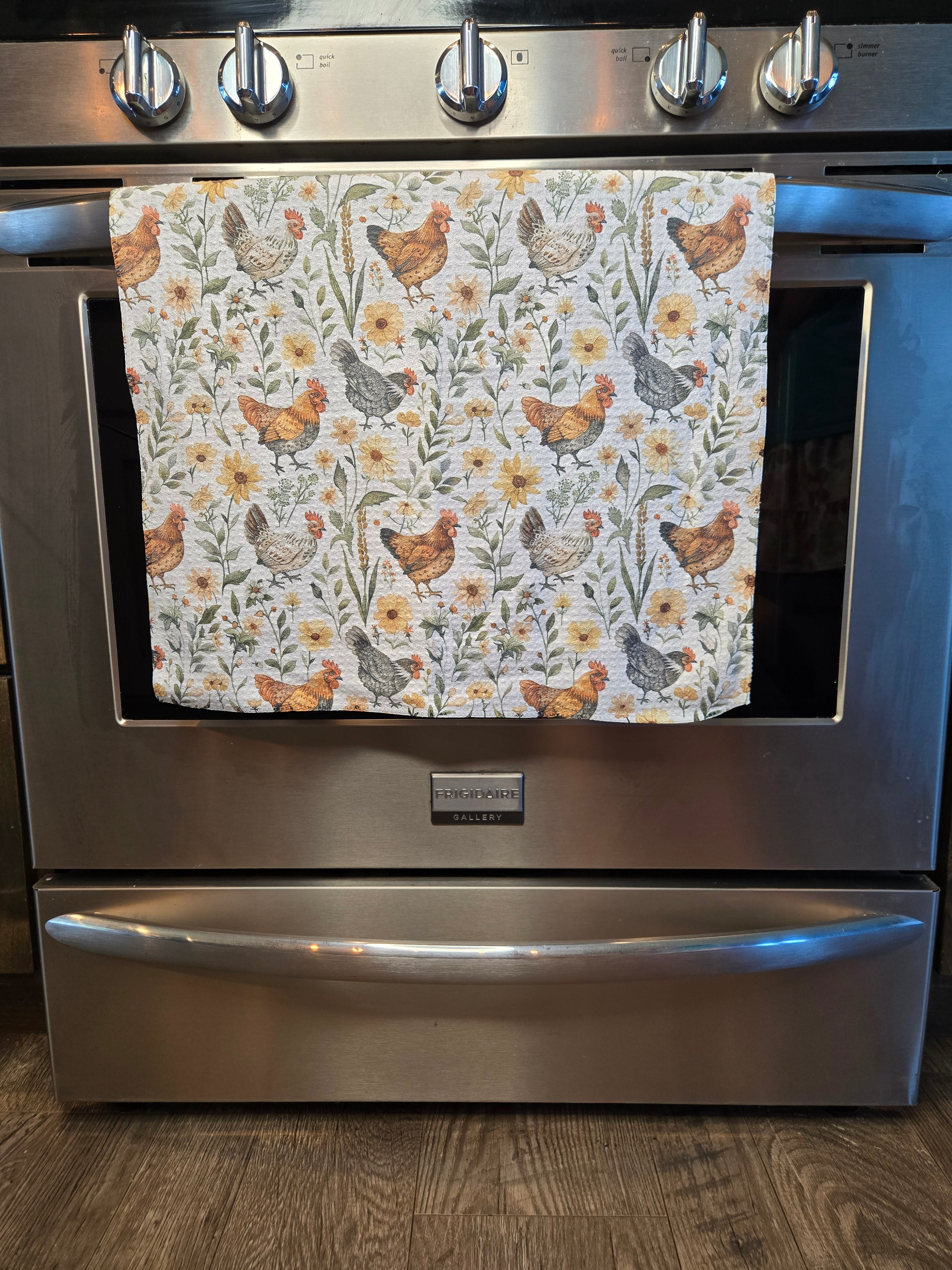 A kitchen towel with a rustic chicken and floral print hanging on the handle of a stainless steel Whirlpool oven.