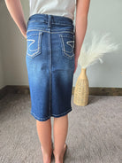 A person shown from the back wearing a blue denim pencil skirt with white contrast stitching and decorative stitching on the back pockets, standing on a gray carpeted floor next to a woven vase with pampas grass.