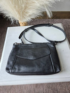 Black leather crossbody handbag with a flap front pocket and adjustable shoulder strap, displayed on a white rectangular surface with dried pampas grass in the background.