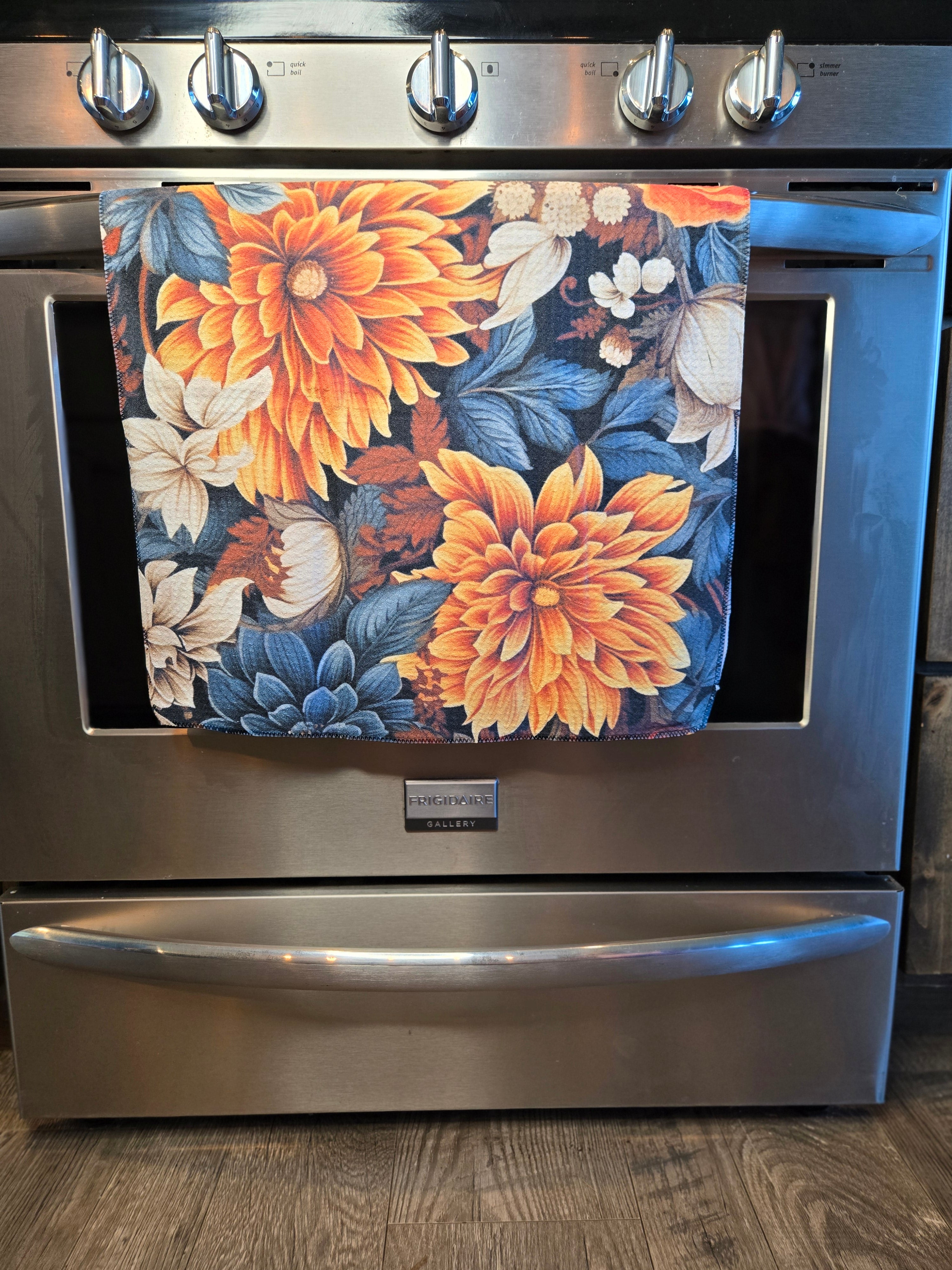 A stainless steel freestanding range oven with a floral kitchen towel hanging from the handle. The towel features large orange and yellow dahlia flowers with blue and white leaves on a dark background.