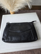 A black leather clutch or wristlet pouch with a front flap pocket, resting on a white wooden board with dried pampas grass in the background.