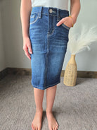 A person wearing a blue denim pencil skirt with contrast stitching, standing barefoot on a gray carpeted floor next to a woven vase with white pampas grass.
