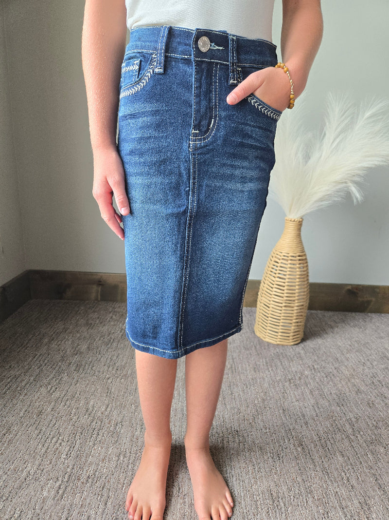 A person wearing a blue denim pencil skirt with contrast stitching, standing barefoot on a gray carpeted floor next to a woven vase with white pampas grass.
