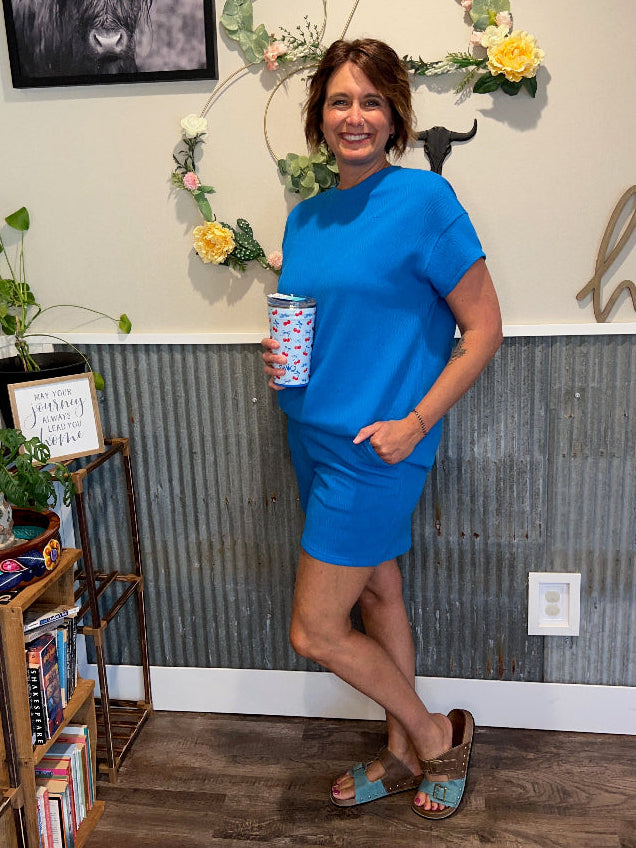 Person in a blue top and shorts set standing in a room with decor elements like plants and a framed picture.