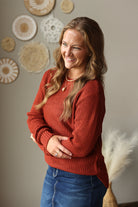 A smiling woman with long wavy light brown hair wearing a rust-orange textured knit sweater and blue jean skirt, standing in front of a neutral wall decorated with circular woven wall art.