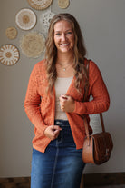 A woman smiling and wearing a rust-orange open-front knit cardigan over a beige top and blue jean skirt, carrying a brown leather shoulder bag. She is standing indoors against a gray wall decorated with circular woven wall hangings.