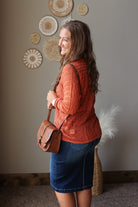 A woman in a rust-orange cable-knit sweater and a blue denim skirt, carrying a brown leather crossbody bag. She is standing in a room with a neutral gray wall decorated with circular woven wall art.