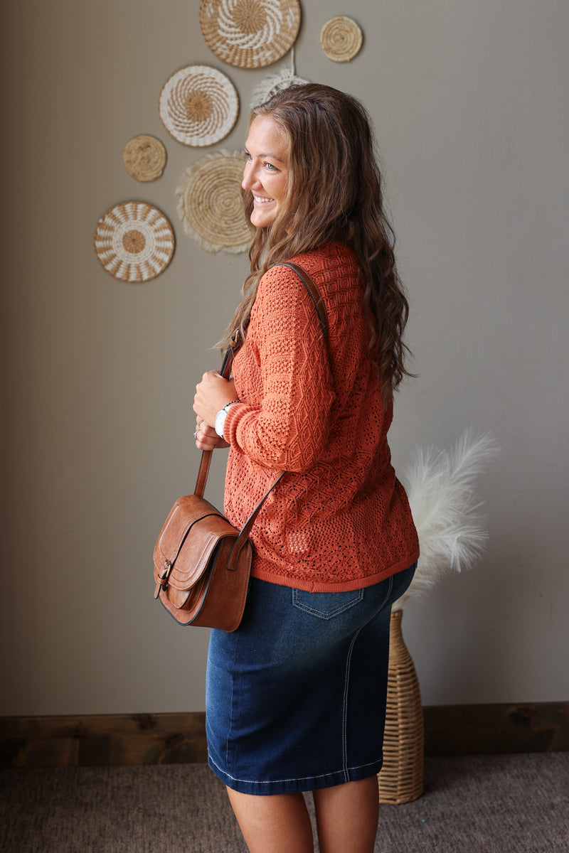 A woman in a rust-orange cable-knit sweater and a blue denim skirt, carrying a brown leather crossbody bag. She is standing in a room with a neutral gray wall decorated with circular woven wall art.