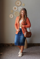 A woman modeling a casual outfit consisting of an orange knit cardigan, a cream tank top, a blue denim midi skirt, and white sneakers, while carrying a brown leather crossbody bag in an indoor setting.