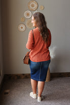 A woman is shown from behind wearing a textured orange knit cardigan, a blue denim pencil skirt, and white slip-on shoes, carrying a brown leather shoulder bag in an indoor setting.