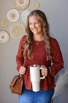 A woman with long wavy light brown hair smiling while holding a white travel mug. She is wearing a rust red long-sleeve blouse with a small yellow and green floral pattern, blue jeans, and carrying a brown leather shoulder bag.