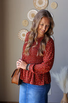 A woman wearing a rust red long-sleeve peplum top with a small white floral pattern, paired with a blue denim skirt. She is holding a brown leather shoulder bag in front of a neutral wall decorated with circular woven wall art.