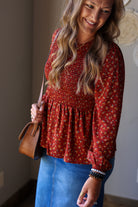 A woman wearing a rust red long-sleeve blouse with a small yellow and white floral pattern and a smocked waist, paired with a blue denim skirt. She is holding a brown leather handbag.