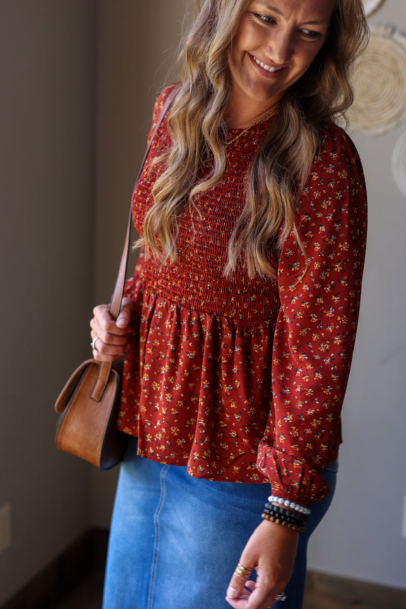 A woman wearing a rust red long-sleeve blouse with a small yellow and white floral pattern and a smocked waist, paired with a blue denim skirt. She is holding a brown leather handbag.