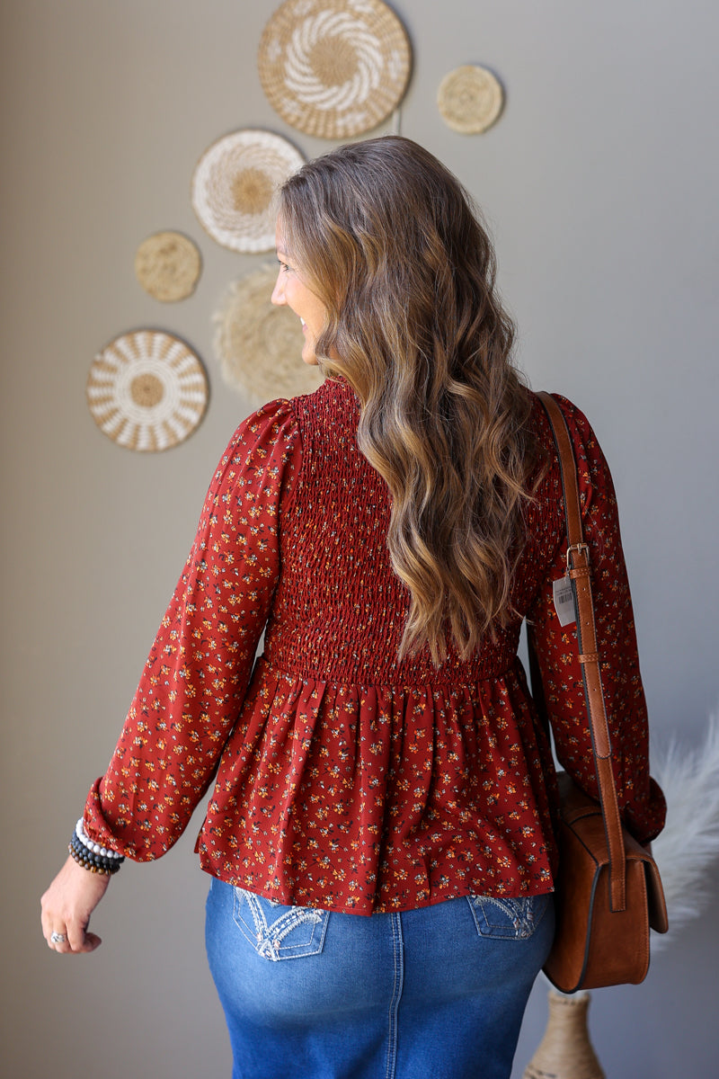 A woman shown from the back wearing a rust red long-sleeve blouse with a small white floral pattern and a smocked waist, paired with blue jeans skirt and a brown shoulder bag.