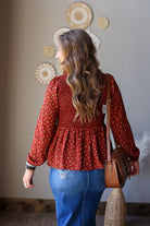 A woman viewed from behind wearing a rust red long-sleeve blouse with a small white floral pattern and a smocked waist, paired with blue jean skirt and a brown leather crossbody bag.