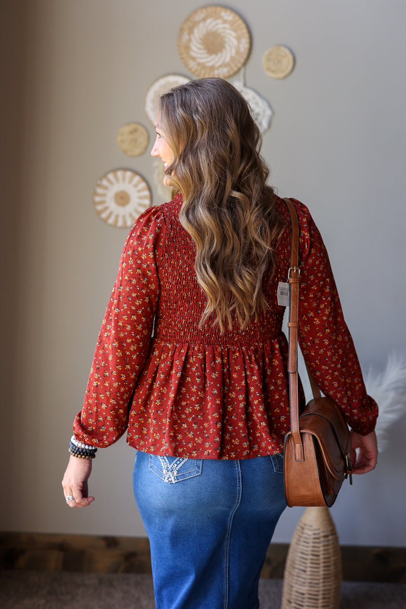 A woman viewed from behind wearing a rust red long-sleeve blouse with a small white floral pattern and a smocked waist, paired with blue jean skirt and a brown leather crossbody bag.