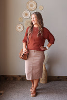 A woman modeling a rust-colored long-sleeve knit top and a khaki taupe pencil skirt, carrying a brown leather crossbody bag. She is standing in a room with a neutral beige wall decorated with circular woven wall art.