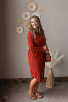 A woman wearing a rust-orange long-sleeve button-up shirt dress, holding a brown leather bag and wearing tan loafers, standing in a styled room with boho decor.