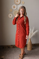 A woman wearing a rust-red corduroy button-down shirt dress, standing in a styled room with a neutral gray wall, decorative wall art, and a woven vase with pampas grass.