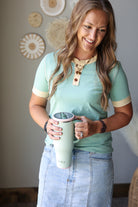A woman wearing a mint green short-sleeve polo shirt with beige trim and a denim skirt holds a matching mint green insulated tumbler with a beige collar detail. She is standing indoors against a neutral wall with decorative circular wall hangings.