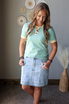 A woman modeling a mint green short-sleeve t-shirt with beige trim and a light blue denim skirt, standing in a room with a gray wall and decorative wall art.