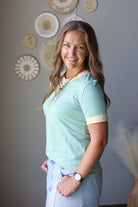 A woman with long brown hair smiling and posing in a mint green short-sleeve polo shirt with beige trim on the collar and sleeve cuffs, paired with light blue jean skirt.