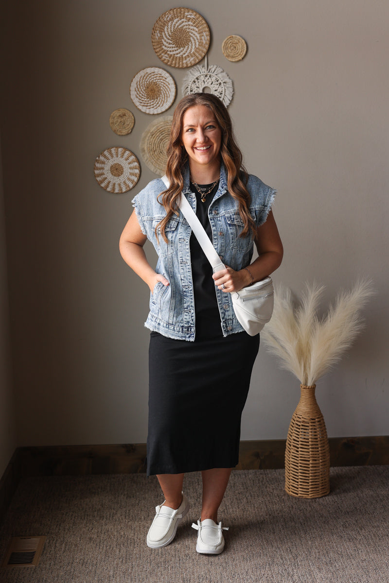 Woman in a denim vest and black dress standing in a room with decorative wall art and a vase.