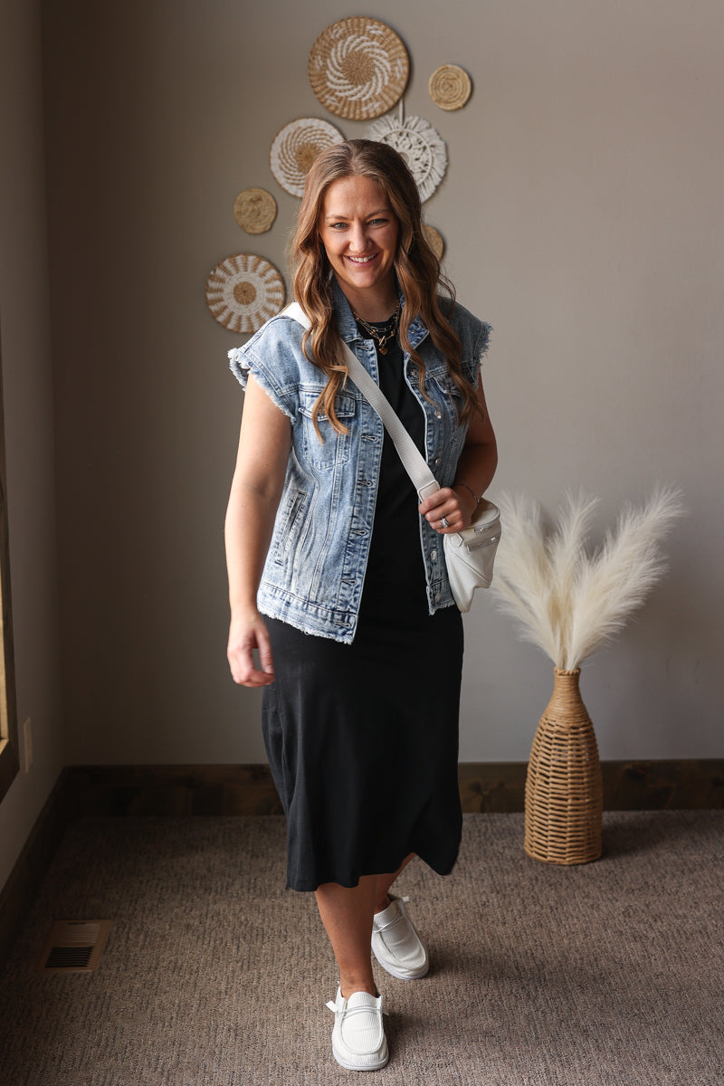 Woman wearing a denim vest over a black dress in a room with decorative wall art and a vase.