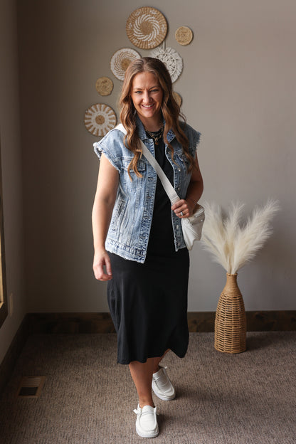 Woman wearing a denim vest over a black dress in a room with decorative wall art and a vase.