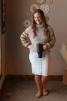 Woman wearing a patterned waffle knit sweater and light wash denim skirt holding a black mug indoors.