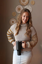 Woman wearing a grey waffle turtleneck sweater with contrast olive print sleeves, holding a black mug in a room with decorative wall art.