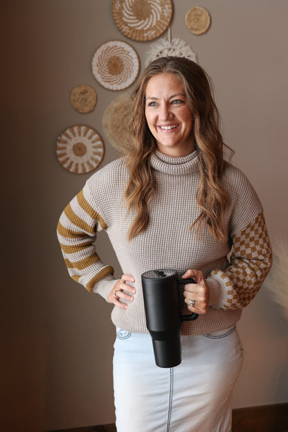 Woman wearing a grey waffle turtleneck sweater with contrast olive print sleeves, holding a black mug in a cozy room with decorative wall art.