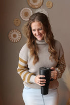 Woman wearing a grey waffle turtleneck sweater with contrast olive print sleeves, holding a black mug indoors with decorative wall art in the background