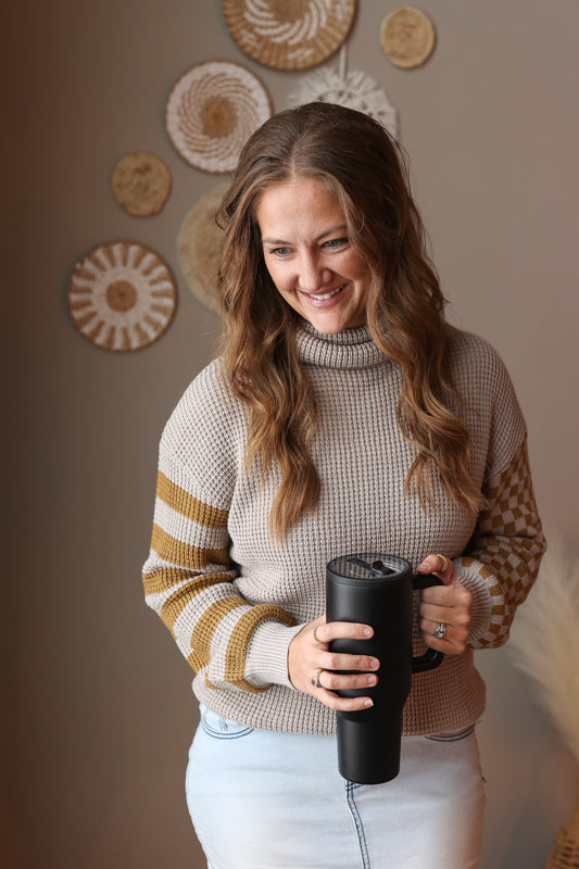 Woman wearing a grey waffle turtleneck sweater with contrast olive print sleeves, holding a black mug indoors with decorative wall art in the background
