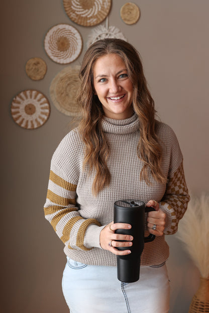 Woman wearing a grey waffle turtleneck sweater with contrast olive print sleeves, holding a black mug in a room with decorative wall art