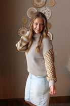 Woman wearing a grey turtleneck sweater with checkered sleeves and a light blue denim skirt, standing against a wall with decorative plates.