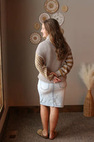Woman wearing a patterned sweater and denim skirt standing in a room with decorative wall hangings.