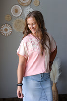 A woman wearing a short-sleeve top with a pink and cream floral lace yoke, paired with a light blue denim skirt, standing in a room with decorative wall art.