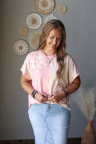 A woman wearing a short-sleeve split-neck blouse with a pink and beige color-blocked design and floral embroidery on the left side, paired with light blue jean skirt, standing in front of a gray wall decorated with circular woven wall art.