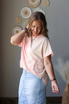 A woman wearing a light peach short-sleeve t-shirt with a pink paisley pattern and light blue denim skirt, standing in a room with decorative wall art.