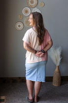A woman stands with her back to the camera wearing a short-sleeve color-block top with cream and dusty pink sections, a light blue skirt, and sandals. The background features a gray wall decorated with circular woven wall art and a tall woven vase with pampas grass.