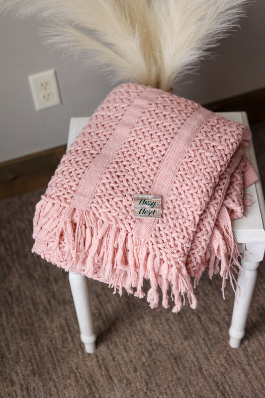 Pink knitted blanket with tassels on a white chair, with a neutral background.