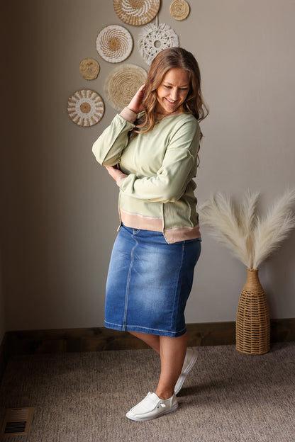 Woman wearing a green sweatshirt and blue denim skirt standing in a room with decorative wall art.