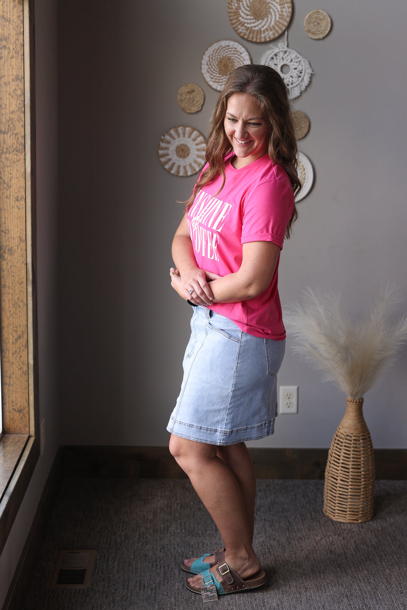 Woman in a pink shirt and denim skirt standing in a room with decorative wall art.