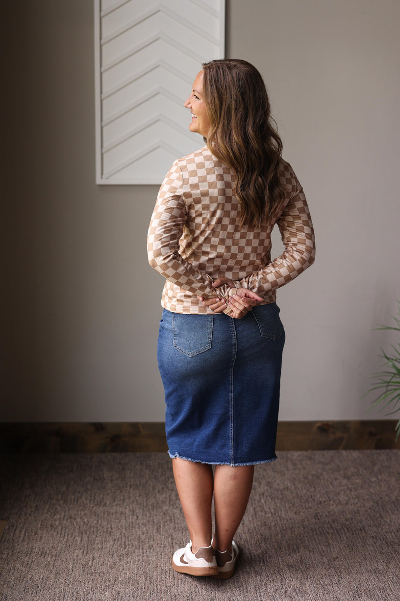 Woman wearing a checkered top and denim skirt standing in a room with a neutral wall and plant.