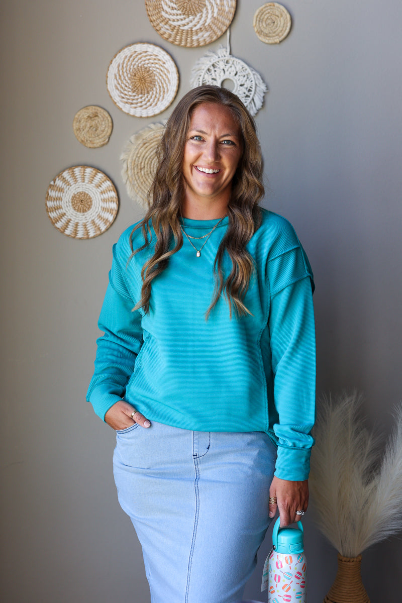 A smiling woman with long wavy brown hair stands indoors wearing a bright turquoise long-sleeve crewneck sweatshirt and light blue jean skirt, holding a patterned water bottle. She is posed against a gray wall decorated with woven wall art.