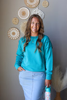 A smiling woman with long wavy brown hair stands indoors wearing a bright turquoise long-sleeve crewneck sweatshirt and light blue jean skirt, holding a patterned water bottle. She is posed against a gray wall decorated with woven wall art.