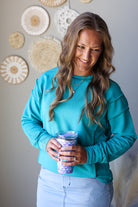 A woman with long wavy brown hair wearing a bright turquoise long-sleeve crewneck sweatshirt and light blue jean skirt, holding a patterned purple and white tumbler cup. She is standing indoors against a light gray wall decorated with circular woven wall art.