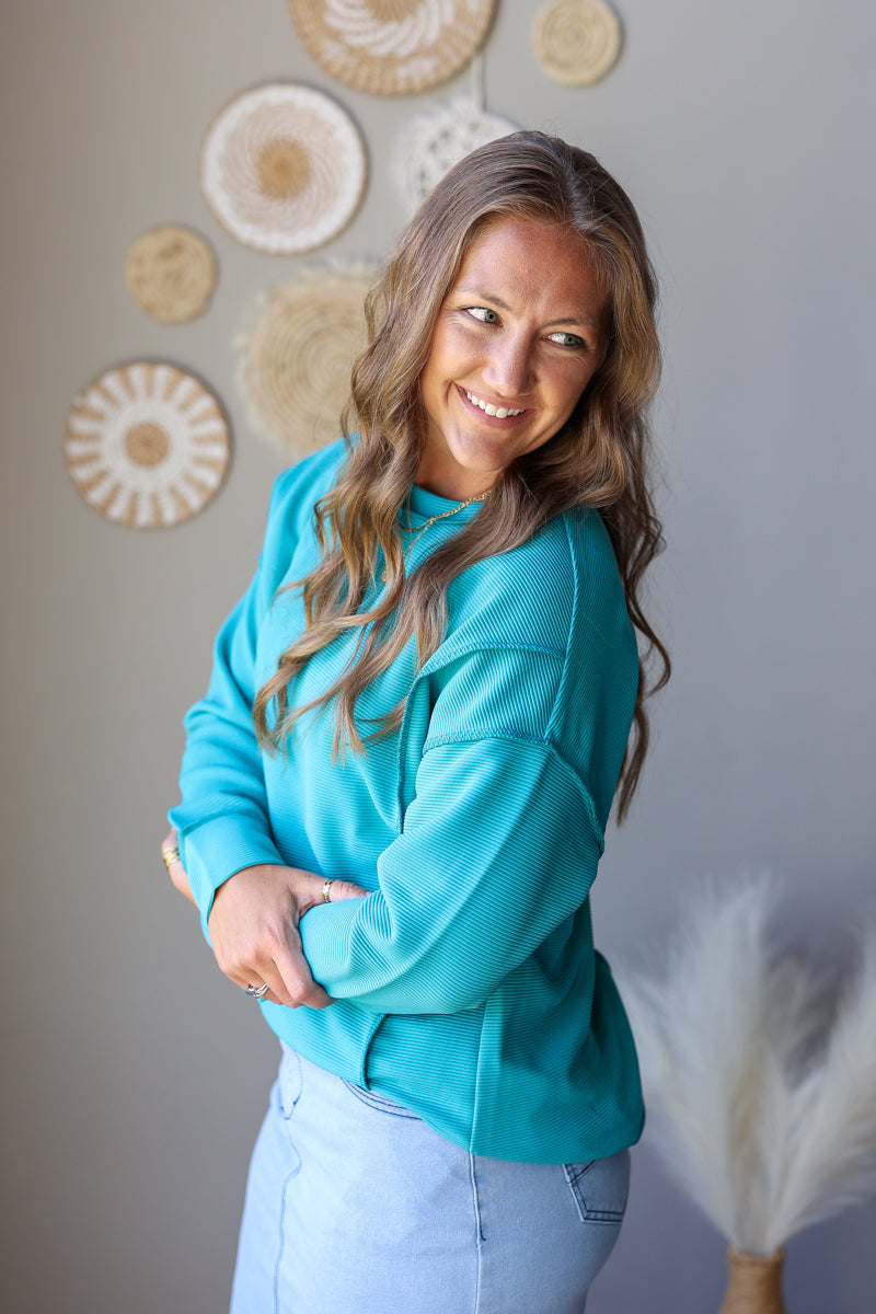 A smiling woman with long wavy brown hair wearing a bright turquoise crewneck sweatshirt and light blue jean skirt, standing against a gray wall decorated with circular woven wall art.